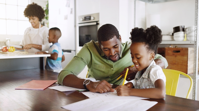 A dad helping a child with homework at the kitchen table and mom and another child in the background. 