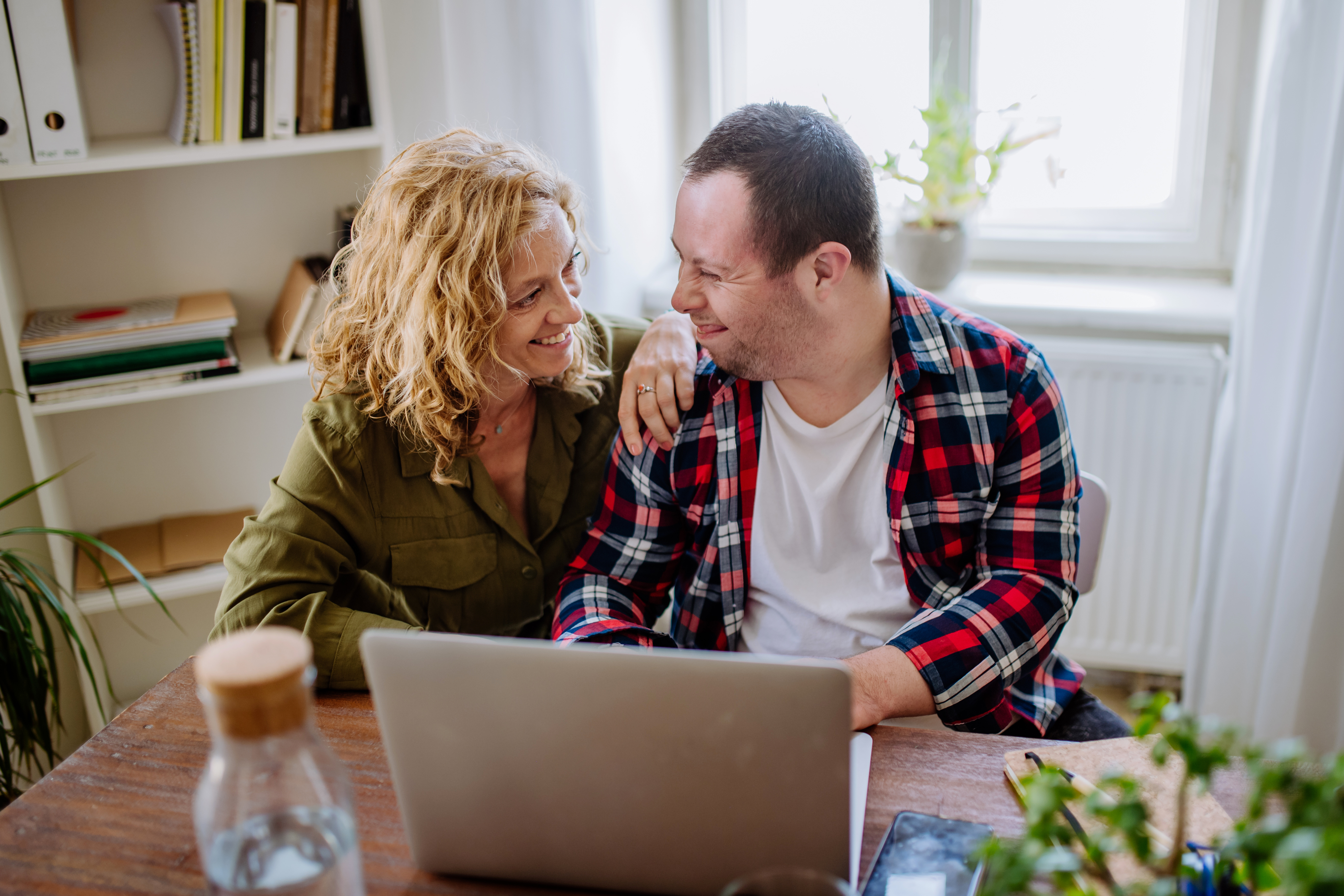 Two people smiling at each other sitting at kitchen table with open laptop.