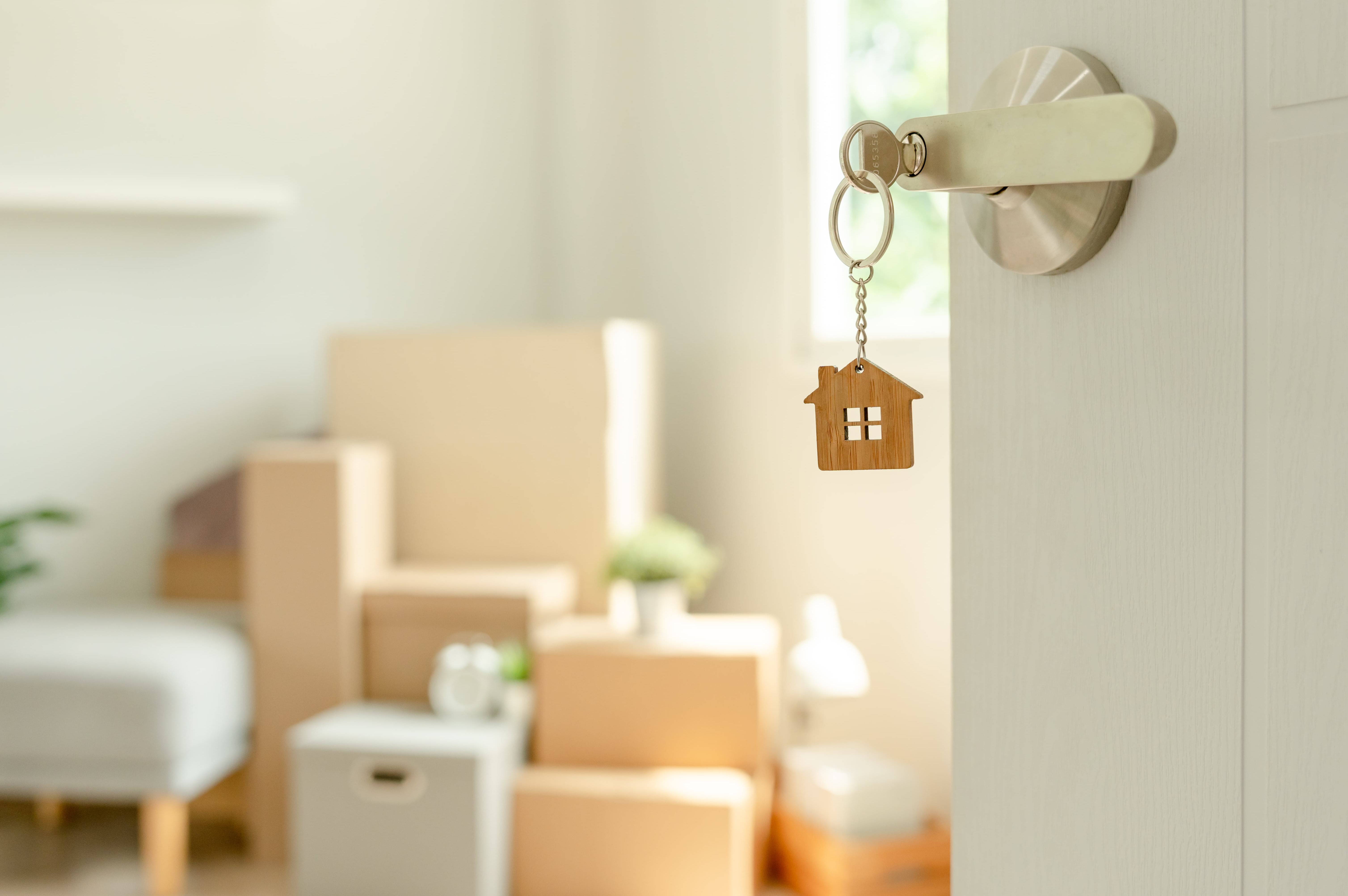 A door slightly open with a key in the lock and a house-shaped keychain, revealing a bright room filled with moving boxes and furniture.
