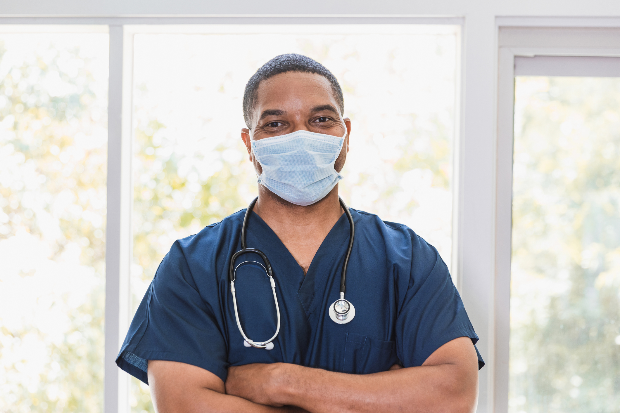 A male nurse wearing a face mask with his arms crossed looks at the camera. 